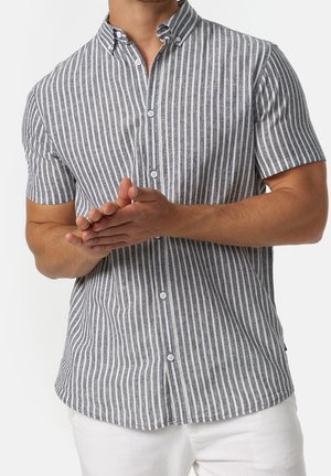 Mann trägt ein graues und weißes, vertikal gestreiftes kurzärmliges Hemd mit weißen Hosen, die Hände auf Taillenhöhe verschränkt.