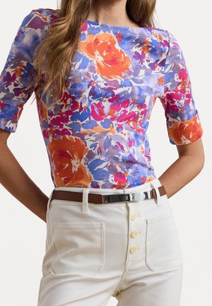 Kvinde iført en tætsiddende bluse med farverige blomsterprint i orange, lilla, pink og blå, kombineret med hvide, højttaljede bukser og et brunt bælte.