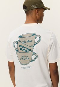 Μπλούζα σε κρεμ χρώμα με γραφικό στοιχείο από στοιβαγμένα φλιτζάνια και μπλε κείμενο «Les Deux SPORTING GOODS» και «Πιείτε μια γουλιά από την καλή ζωή.»