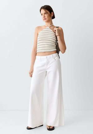Joven mujer con pantalones blancos de pierna ancha y top de tirantes a rayas, sosteniendo un bolso de hombro sobre un fondo liso.