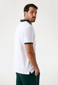 Weißes Poloshirt mit grünem Kragen und Ärmeln, ausgestattet mit einem seitlichen Schlitz. Der Stoff wirkt glatt und atmungsaktiv.
