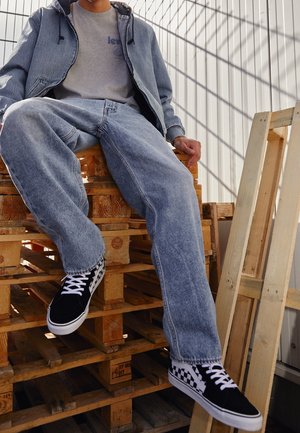 Persona vistiendo chaqueta vaquera azul claro y vaqueros con zapatillas de cuadros en blanco y negro, sentada sobre palets de madera apilados.