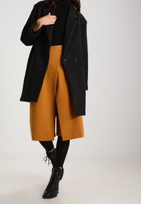 Donna con un cappotto nero, dolcevita nera, culotte a gamba larga senape, collant neri e stivaletti neri con lacci su uno sfondo neutro.