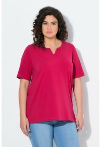 Top a manica corta a V in cotone rosa brillante, vestibilità ampia. Presenta un sottile dettaglio di cucitura lungo il colletto, abbinato a jeans azzurri chiari.
