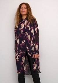 Camicia lunga e trasparente con un motivo astratto in viola, bianco e nero, dotata di una chiusura frontale con bottoni e un orlo curvo. Abbinata a leggings neri.