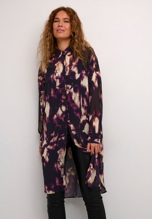 Camicia lunga e trasparente con un motivo astratto in viola, bianco e nero, dotata di una chiusura frontale con bottoni e un orlo curvo. Abbinata a leggings neri.