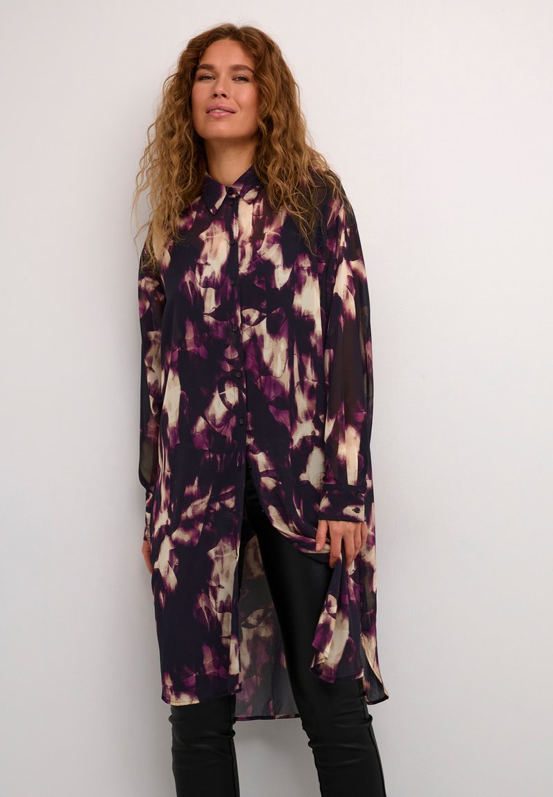 Camicia lunga e trasparente con un motivo astratto in viola, bianco e nero, dotata di una chiusura frontale con bottoni e un orlo curvo. Abbinata a leggings neri.