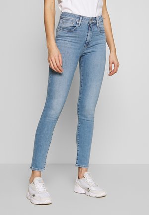 Personne portant un jean skinny bleu clair, un t-shirt blanc et des baskets blanches, debout devant un fond gris uni.