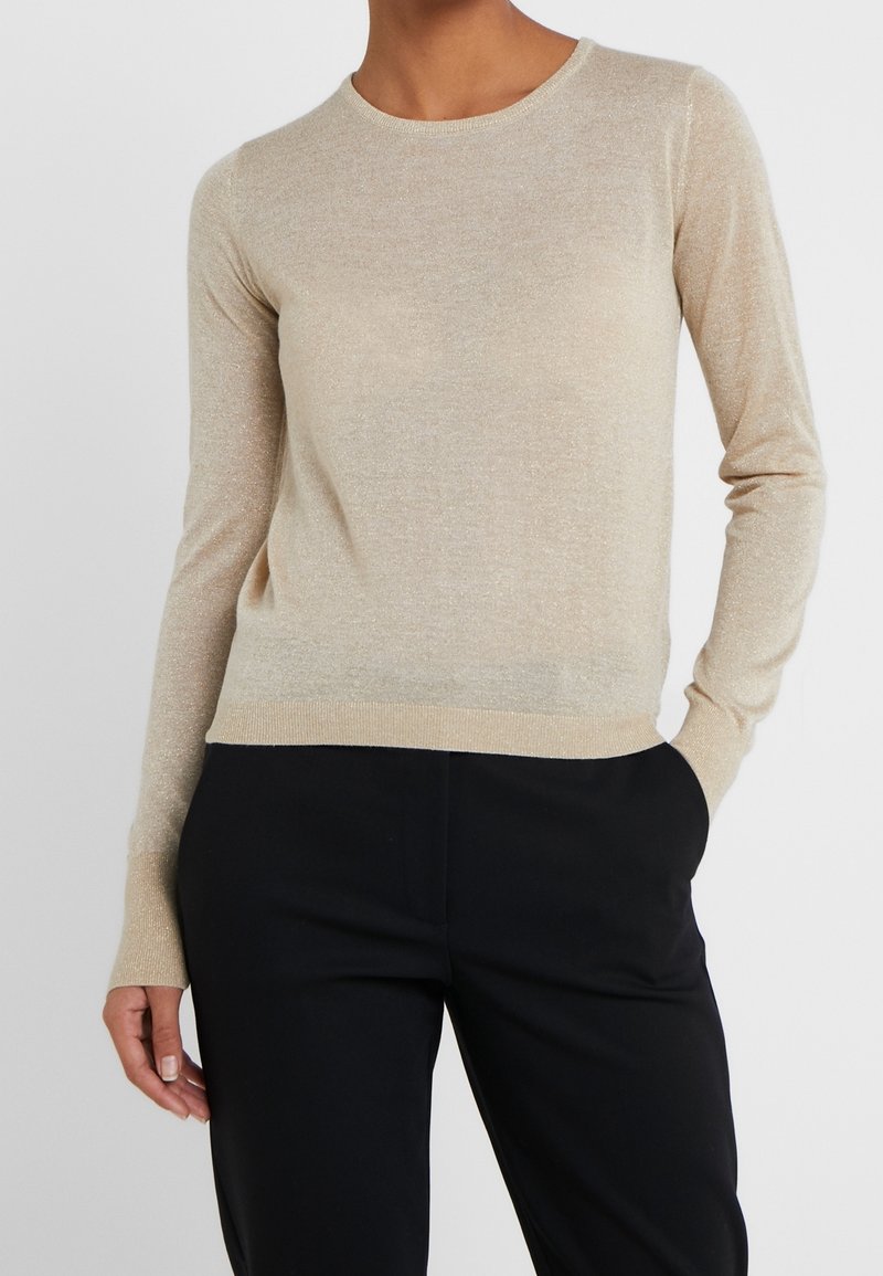 Maglione beige a maniche lunghe realizzato in tessuto scintillante, con scollatura a girocollo e design aderente, abbinato a pantaloni neri. Stile elegante e minimalista.