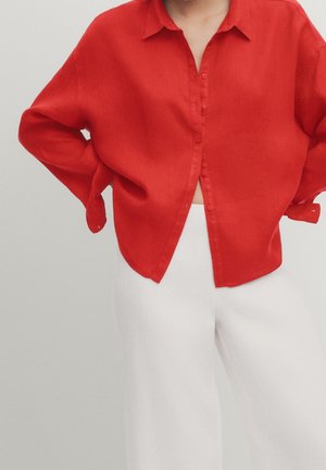 Osoba ubranа w luźną czerwoną koszulę na guziki, z rękoma na biodrach, w białych spodniach z wysokim stanem na jednolitym tle.