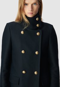 Abrigo marinero de doble botonadura en tejido texturizado con botones dorados, cuello alto y bolsillos laterales, con una silueta ajustada.