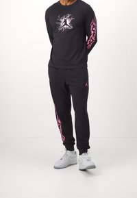 Schwarzes langärmliges Hemd und Jogginghosen mit pinken Grafiken. Das Hemd hat ein zentrales Logo; die Hose hat seitliche Streifendetails. Weiße Sneaker.