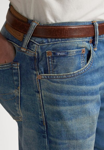 Tuva zila džinsa bikses ar Polo zīmola etiķeti, valkātas kopā ar brūnu ādas jostu un roku kabatā, kombinētas ar baltu kreklu.