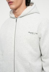 Haut de survêtement à zip gris clair avec une capuche doublée en sherpa et des poches avant. Présente une broderie de marque sur la poitrine et un tissu texturé.