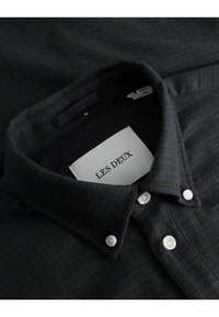 Chemise noire texturée avec col boutonné, arborant une étiquette "LES DEUX" et des boutons blancs. Motif rayé subtil dans des tons sombres.