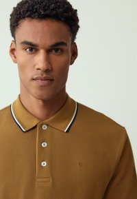 Camisa polo marrón con un tejido texturizado, que presenta una tapeta de tres botones y un cuello con acentos en azul y blanco. Logo sutil bordado en el pecho.