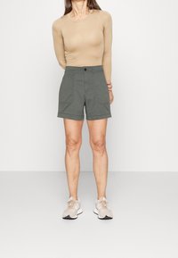 Shorts de color verde oliva fabricados en algodón con cierre de botón, presentan bolsillos laterales y un corte relajado. Combinados con una blusa de manga larga beige y zapatillas ligeras.