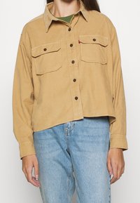 Camicia in corduroy marrone chiaro con maniche lunghe, due tasche sul petto e bottoni marroni. Abbinata a jeans azzurri sbiaditi.
