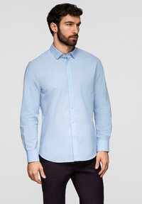 Homme aux cheveux foncés et barbe, portant une chemise à manches longues bleu clair boutonnée et un pantalon foncé, debout devant un fond clair uni.