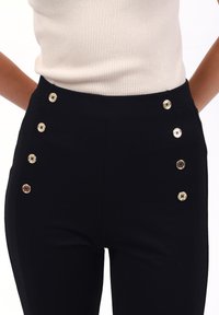 Pantaloni neri a vita alta con una texture liscia, dotati di sei bottoni decorativi dorati sul lato per un tocco in più.
