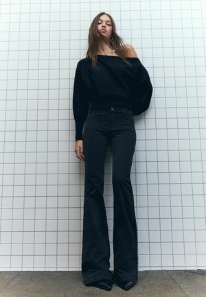 Black off-shoulder sweater with ribbed waistband, paired with fitted black flared jeans. High-heeled black shoes complete the look.