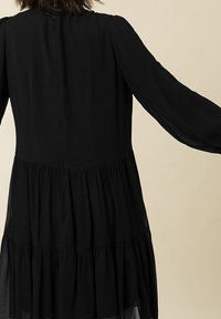 Mustapuku pitkillä hihoilla, kerroksellinen muotoilu ja läpikuultava kangas. Mukana pyöreä pääntie ja takana vetoketjukiinnitys, joka luo virtaavan siluetin.