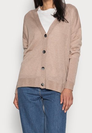 Femme portant un cardigan beige boutonné, une chemise blanche en dessous et un jean bleu, debout devant un fond clair uni.