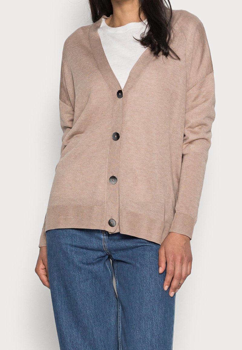 Femme portant un cardigan beige boutonné, une chemise blanche en dessous et un jean bleu, debout devant un fond clair uni.