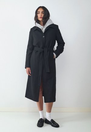 Junge Frau trägt einen schwarzen, taillierten Mantel über einem grauen Hoodie, weiße Socken und schwarze Loafers, steht vor einem schlichten weißen Hintergrund.