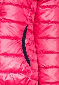 Doudoune rose avec un design matelassé, une texture brillante, et des poches latérales avec des accents en tissu bleu marine. Légère et résistante à l'eau.