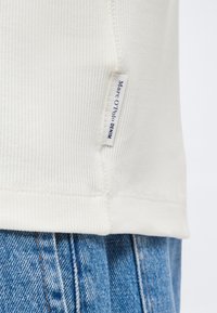 Κοντινό πλάνο του ραβδωτού λευκού υφάσματος στο τελείωμα πουκάμισου με μια μικρή λευκή ετικέτα που γράφει "Marc O'Polo DENIM" πάνω από μπλε τζιν τζιν.