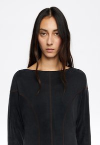 Top negro de manga larga con un escote amplio, que presenta costuras marrones en contraste, textura suave y elementos de diseño estructurados.