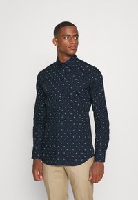 Camicia a maniche lunghe blu navy con pois bianchi, chiusura con bottoni frontale e colletto regolare. Realizzata in tessuto liscio con un taglio aderente, presenta bottoni a contrasto.