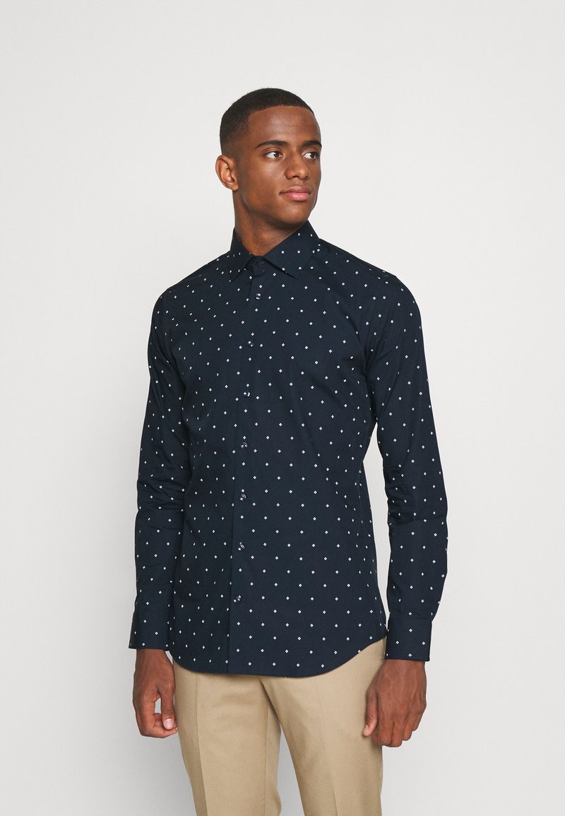 Camicia a maniche lunghe blu navy con pois bianchi, chiusura con bottoni frontale e colletto regolare. Realizzata in tessuto liscio con un taglio aderente, presenta bottoni a contrasto.