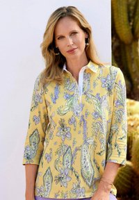 Langarmige, met kraag, gele blouse met een bloemen- en paisleypatroon. Heeft een witte sluiting en een zachte, lichte textuur.