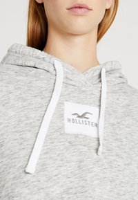Šedá měkká mikina s kapucí s texturovaným povrchem, bílými šňůrkami a přední nášivkou se stříbrným logem "Hollister".