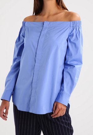 Vrouw draagt lichtblauwe off-shoulder blouse met lange mouwen en zwarte pantalon met fijne strepen tegen een effen achtergrond.
