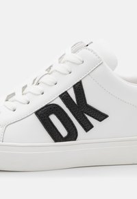 Hvit sneaker med svart teksturert "DK" bokstaver, med runde lisser, glatt læroverdel og gummisåle. Minimalistisk design.