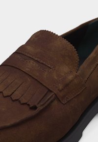 Καφέ δερμάτινο μοκασίνι από σουέτ με διακοσμητικά φουντάκια και μια διακοσμητική λουρίδα στην κορυφή, που δείχνει υφασμένη ραφή και κυμματιστές άκρες.