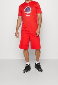 Conjunto atlético rojo que consta de una camiseta de manga corta con un logo circular y pantalones cortos a juego. Zapatillas deportivas negras con detalles en blanco.