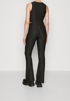 Femme portant un top sans manches noir à fines rayures et un pantalon évasé assorti avec des chaussures plateformes noires épaisses, debout contre un mur uni.