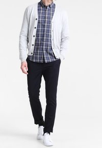 Ljusgrå cardigan över en blå rutad skjorta, i kombination med mörka byxor och vita sneakers. Cardigan har knappar framtill.