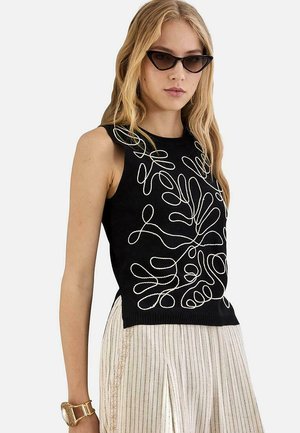 Mujer con top sin mangas negro con diseño abstracto de líneas blancas, pantalones claros a rayas, gafas de sol estilo cat-eye y pulsera dorada.