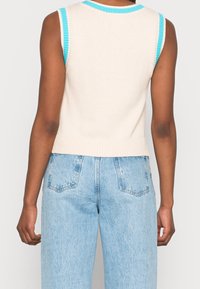 Beige stickad ärmlös topp med turkosa ribbade ringningar och ärmhål. Korthåret design med en texturerad yta. Kombineras med ljusblå hög midja jeans.