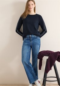 Femme portant un t-shirt à manches longues bleu marine et un jean bleu, debout à côté d'un tabouret sur lequel est posé un sweat polaire violet plié.