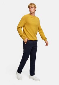 Pull tricoté jaune moutarde avec poignets et encolure côtelés, associé à un pantalon bleu marine foncé et des baskets blanches, présentant une coupe décontractée.