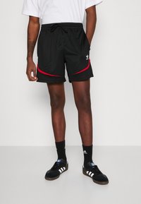 Pantaloni sportivi neri con dettagli curvi rossi, vita elastica e tasche laterali. Abbinati a sneakers nere Adidas e calze alla caviglia.