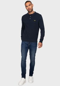 Námořnický pletený polo svetr s texturovaným designem, s límcem a malým žlutým logem, v kombinaci s slim-fit modrými džíny a bílými teniskami.