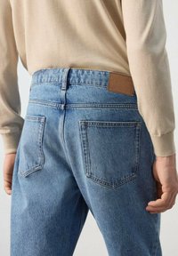 Persona vistiendo jeans de mezclilla azul claro y una camisa de manga larga beige, mostrada desde la cintura hacia abajo, con las manos relajadas a los lados.