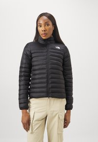 Giacca a vento nera con colletto alto, motivo trapuntato orizzontale e zip frontale. Il logo The North Face è visibile sul lato sinistro del petto.